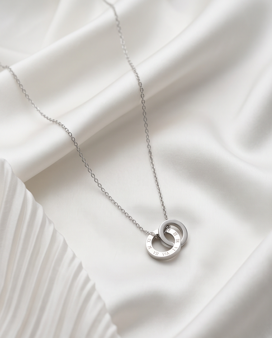 The Roman interlocked Roman numeral circle necklace in silver PVD surgical steel on white fabric, Stacked by Jayneebell