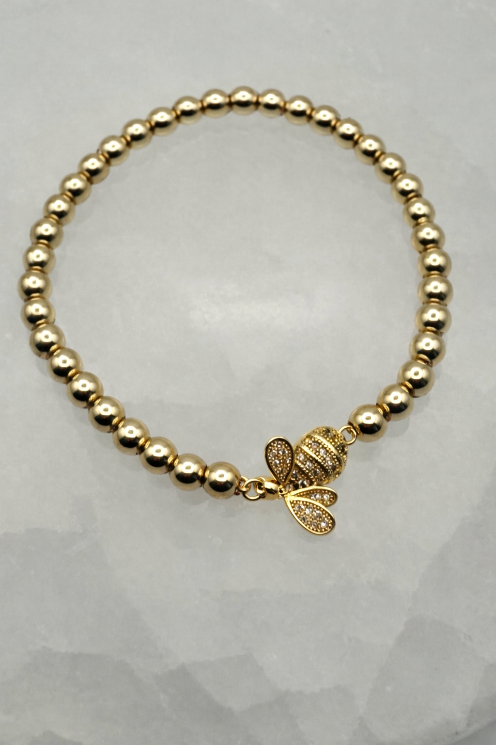 Gold beaded bracelet with a gold bee charm on a light gray background