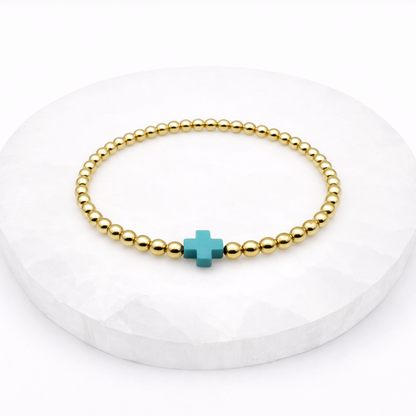 Gold beaded bracelet with a turquoise cross charm on a white background