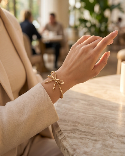 Gold filled bow bracelet worn on wrist in restaurant setting — The Coquette by Stacked by Jayneebell