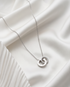 The Roman interlocked Roman numeral circle necklace in silver PVD surgical steel on white fabric, Stacked by Jayneebell