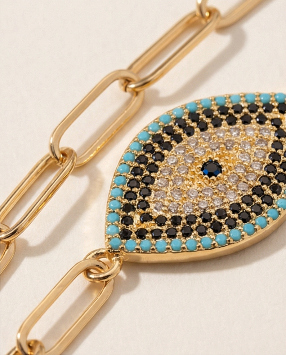 Gold chain bracelet with an eye-shaped charm featuring turquoise, black, and clear gemstones on a beige background.