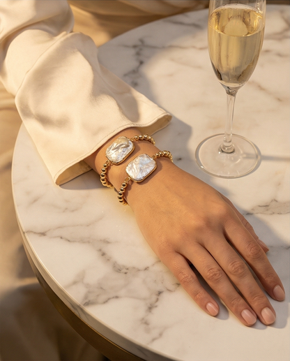 Hand wearing a gold bracelet with heart-shaped gemstones on a marble surface with a glass of champagne.