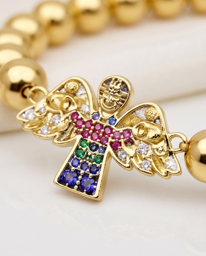 Gold bracelet with a colorful gemstone cross charm on a light background. 14k gold plated angel charm stretch bracelet with multicolor CZ flat lay — Heaven Sent by Stacked by Jayneebell